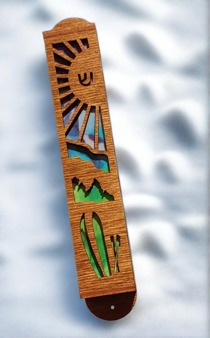 Honey brown mezuzah with cut out image of sun, mountains, and skis. A green and blue background is showing through the cut outs.