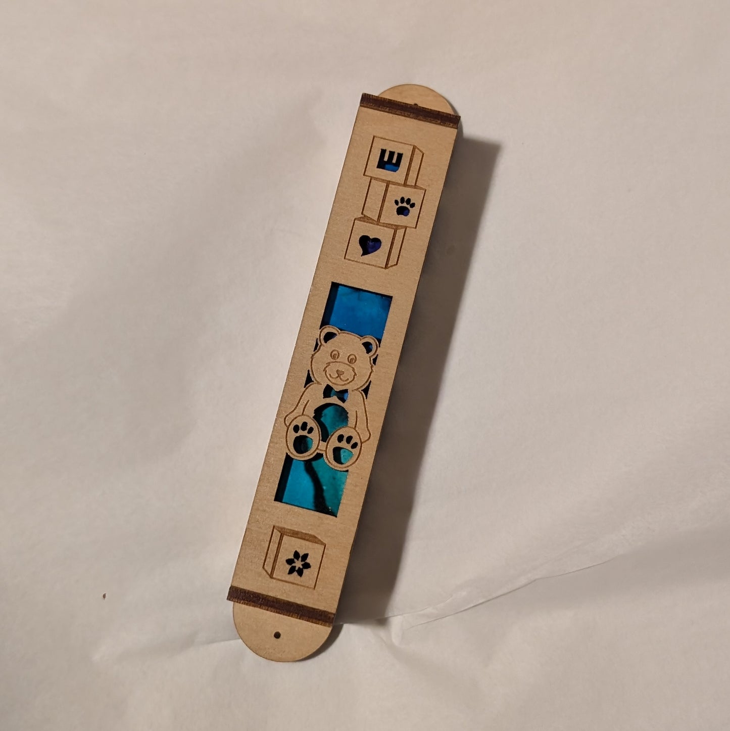 Wooden laser cut mezuzah case decorated with a teddy bear and play blocks. Blue-green background showing through.