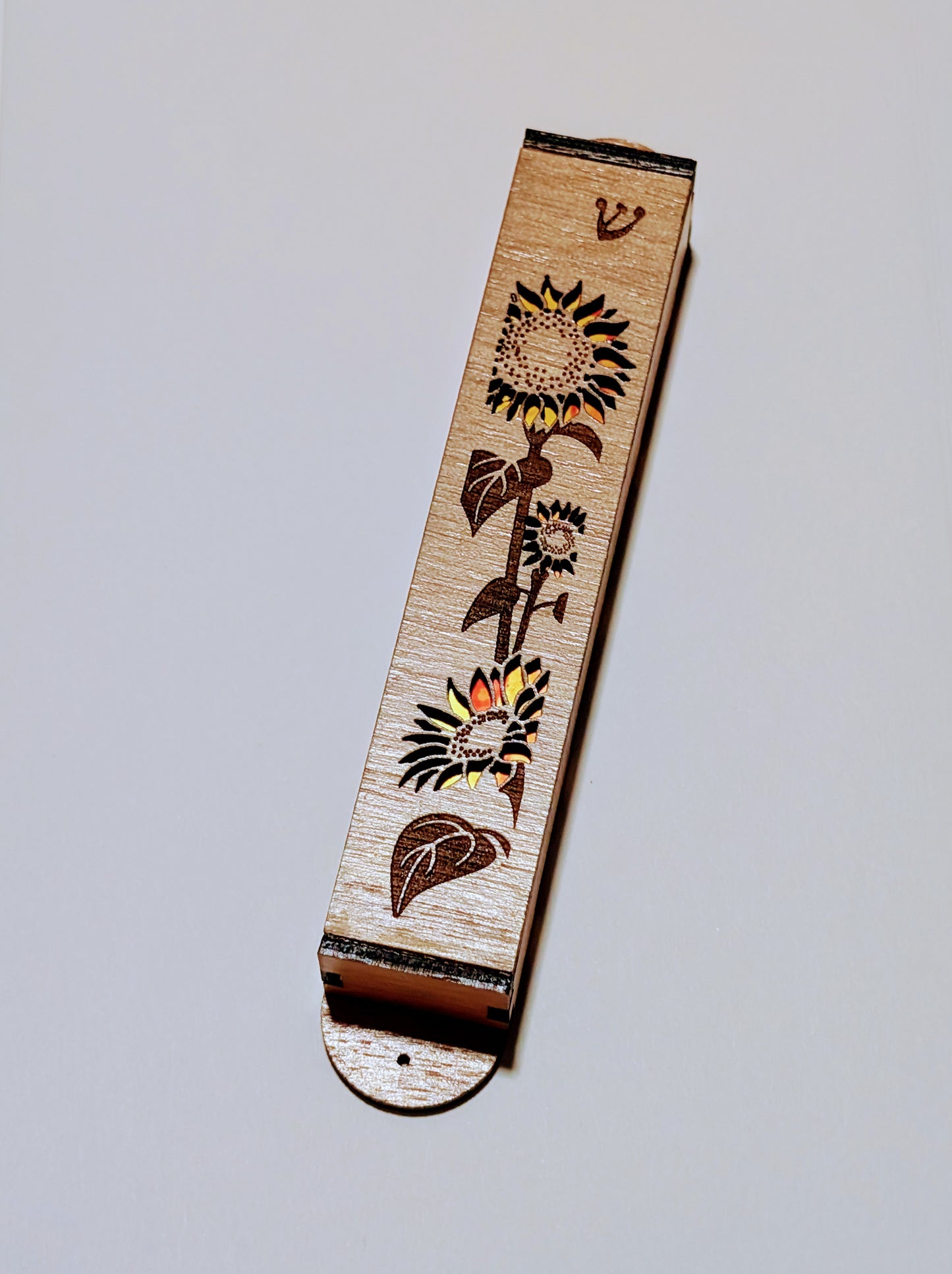 A wooden mezuzah case with a sunflower engraved onto the surface and the petals are cutout with yellow and orange showing through.