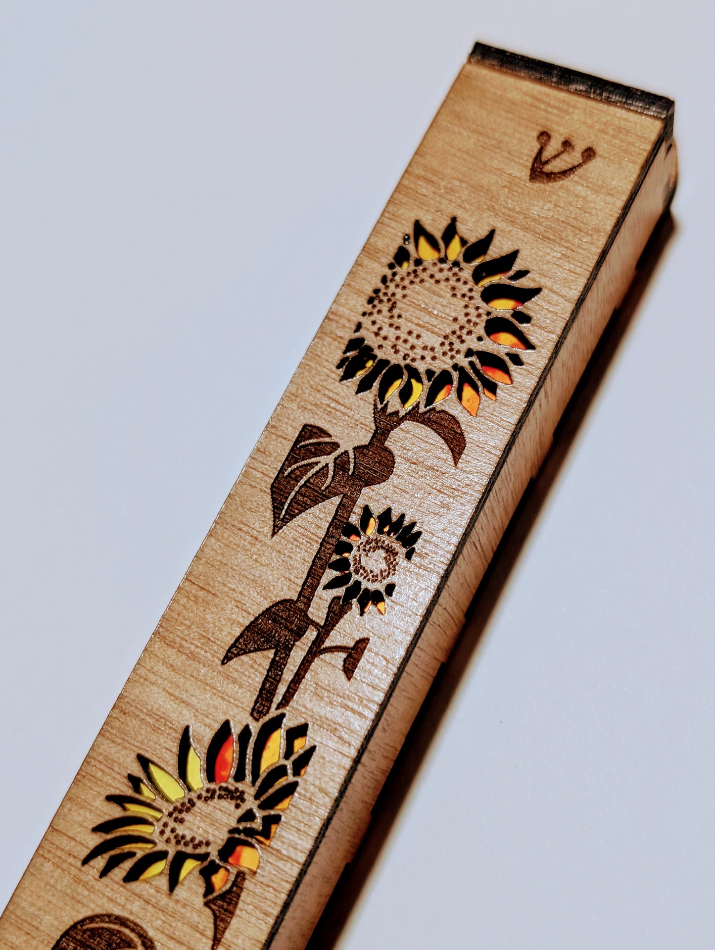 A wooden mezuzah case with a sunflower engraved onto the surface and the petals are cutout with yellow and orange showing through.