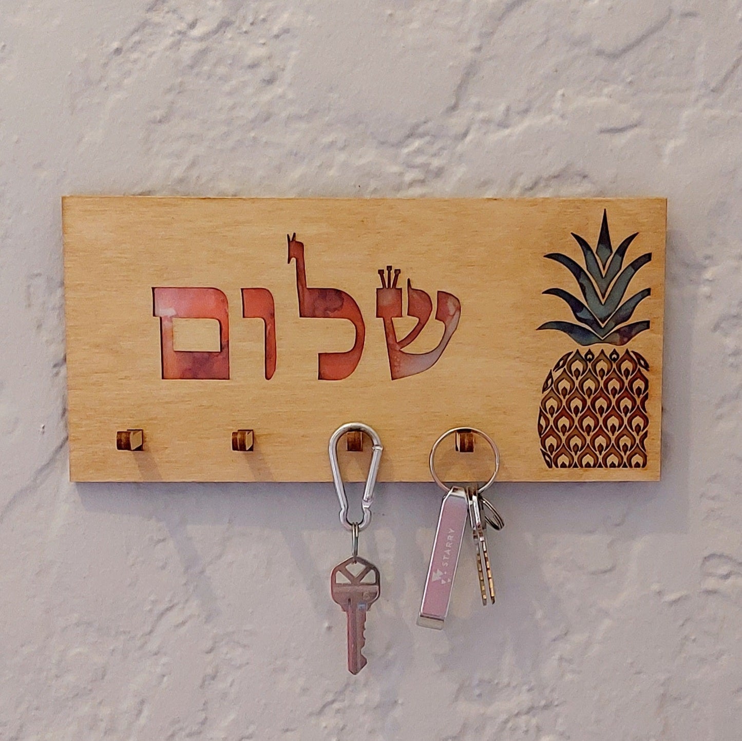Lightly stained wood rectangular key holder. Cutout "שלום" with red background, pineapple with green, yellow, and orange, and 4 pegs for keys with keys hanging on them.