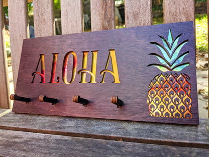 Dark stained wood rectangular key holder. Cutout "Aloha" with yellow and orange background,pineapple with green, yellow, and orange, and 4 pegs for keys.