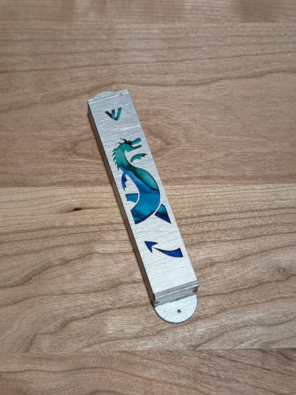 Silver painted mezuzah case with the cutout of a dragon and blue painted background showing through.