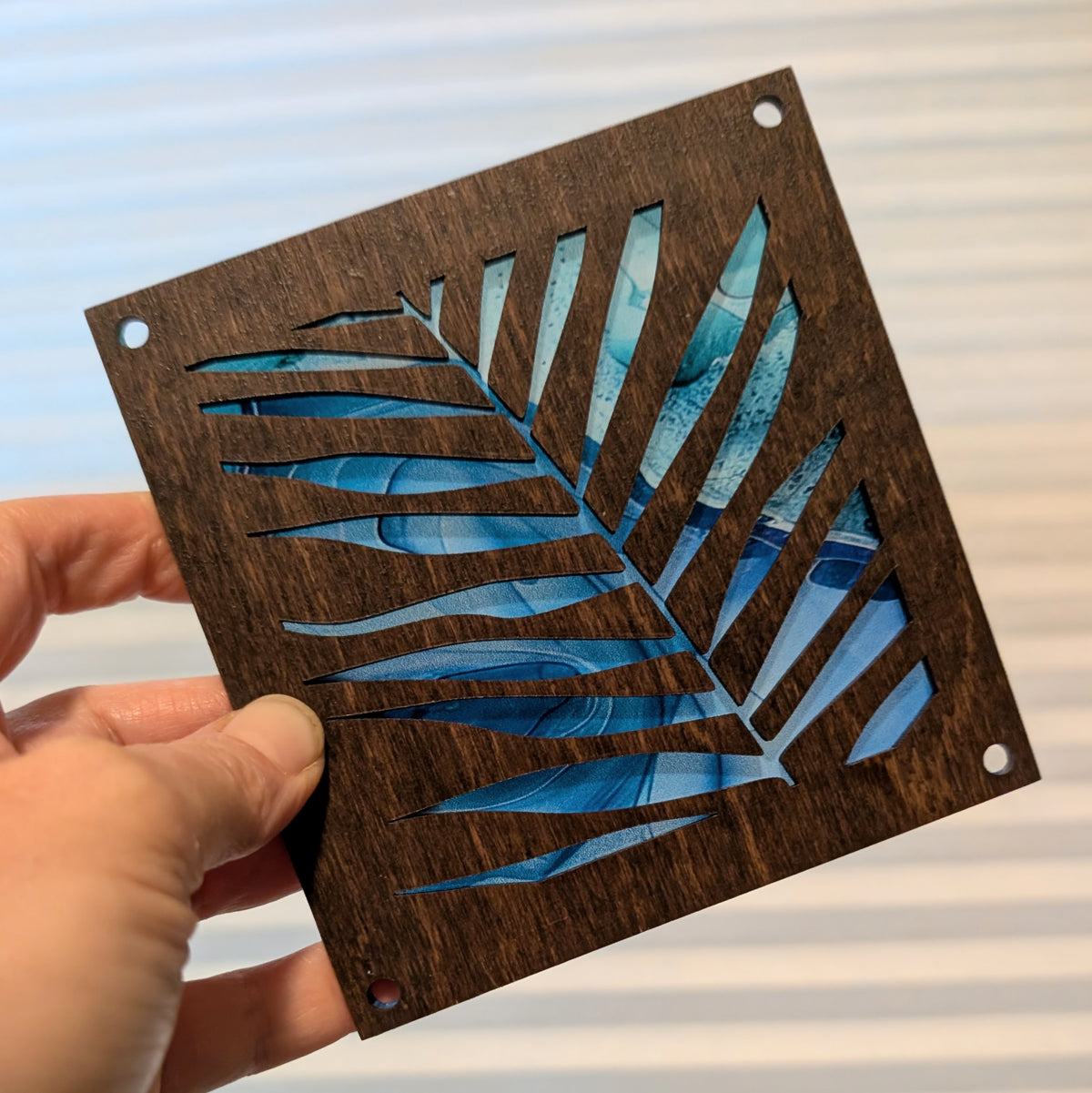 A hand holds a square dark brown wooden plaque featuring a laser-cut tropical palm leaf design with blue and light blue marbled background. The plaque has pre-drilled corner holes and is photographed against a soft-focus background of horizontal window blinds.