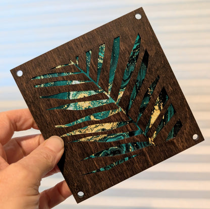 A hand holds a square dark brown wooden plaque featuring a cutout tropical palm leaf design filled with teal and white marbled inlay. The plaque has pre-drilled holes.