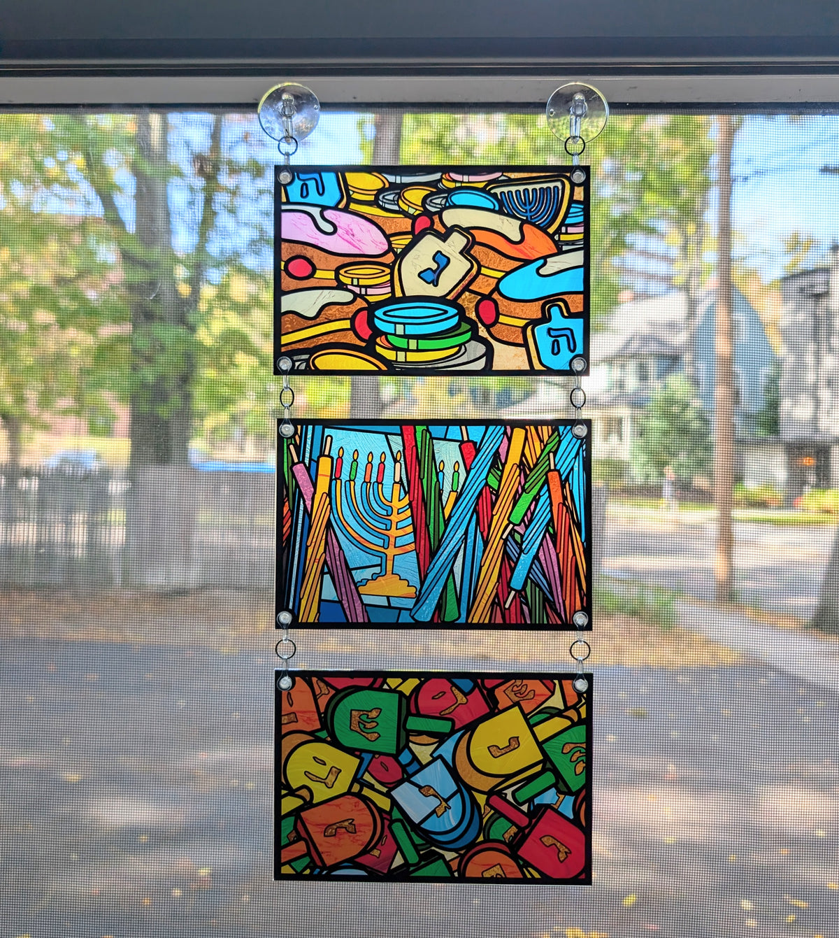 3 panels hanging in a window. Stained glass style art with Hanukah food, candles, and dreidels 