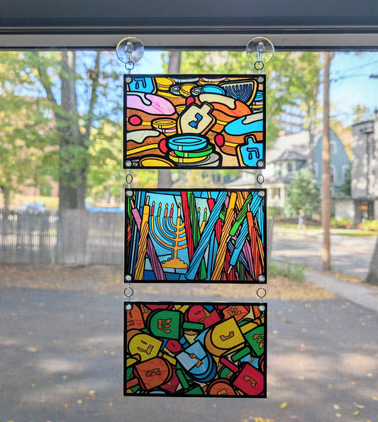 3 panels hanging in a window. Stained glass style art with Hanukah food, candles, and dreidels 
