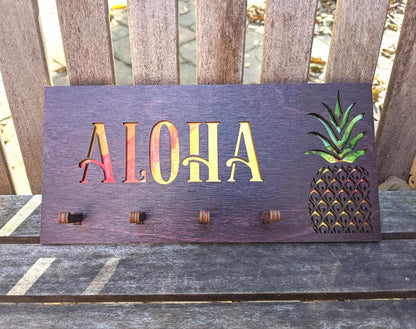 Dark Stain wood rectangle with the word, "Aloha" and a pineapple cut out with yellow, orange, and green showing through. 4 pegs for keys.