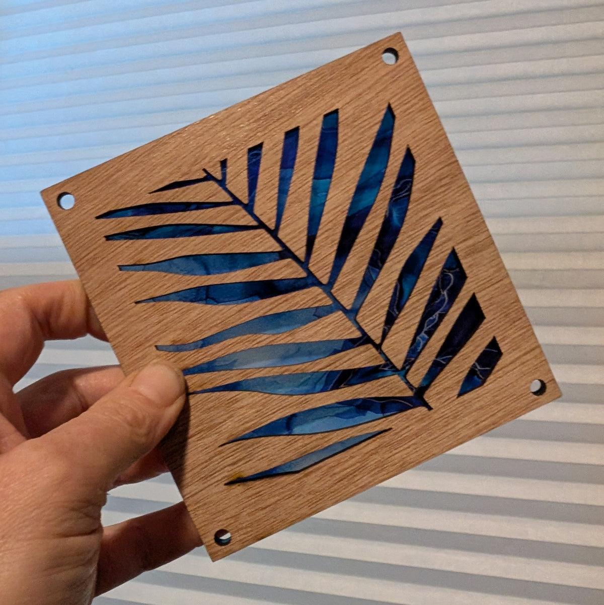 A hand holds a square wooden plaque featuring a cutout palm leaf design with a blue swirled background, set against a background of white window blinds.