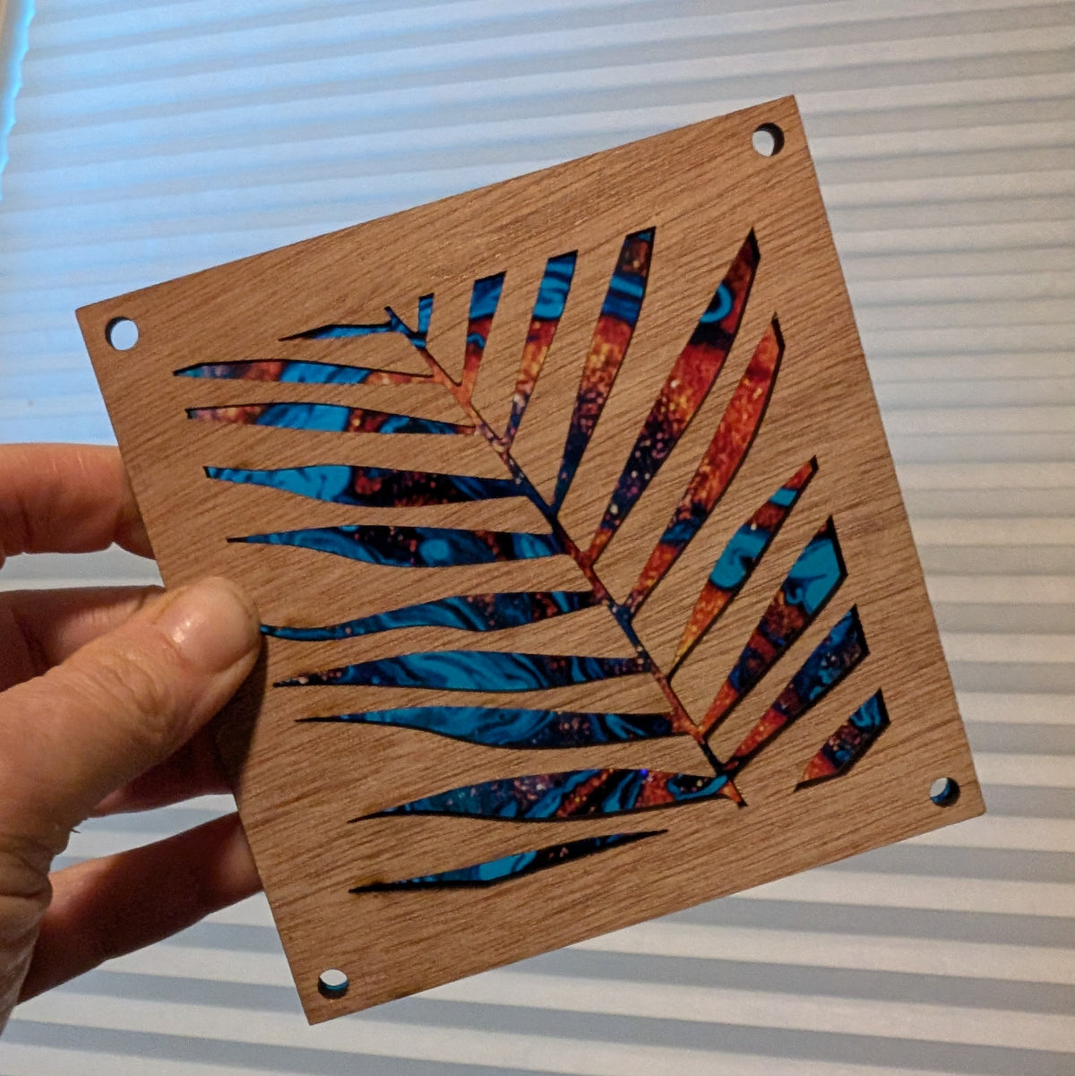 A hand holds a square wooden square featuring a laser-cut palm leaf design with colorful patterned inlay. The piece has pre-drilled holes and is shown against a background of white window blinds.