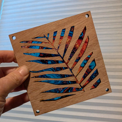 A hand holds a square wooden square featuring a laser-cut palm leaf design with colorful patterned inlay. The piece has pre-drilled holes and is shown against a background of white window blinds.