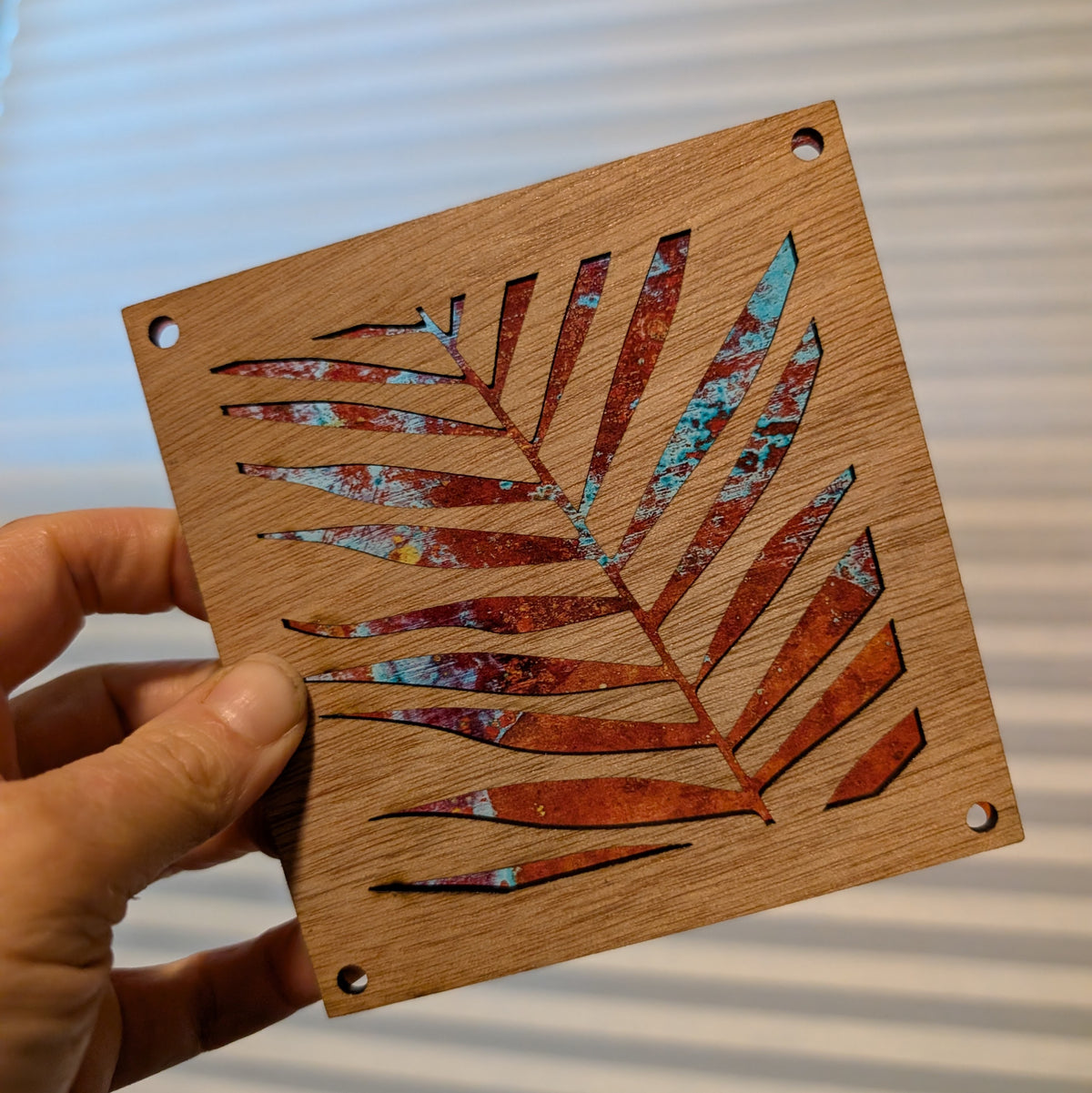 A hand holds a square wooden piece featuring a laser-cut palm leaf design with a distressed copper and turquoise inlay over a natural wood grain background.