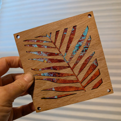 A hand holds a square wooden piece featuring a laser-cut palm leaf design with a distressed copper and turquoise inlay over a natural wood grain background.
