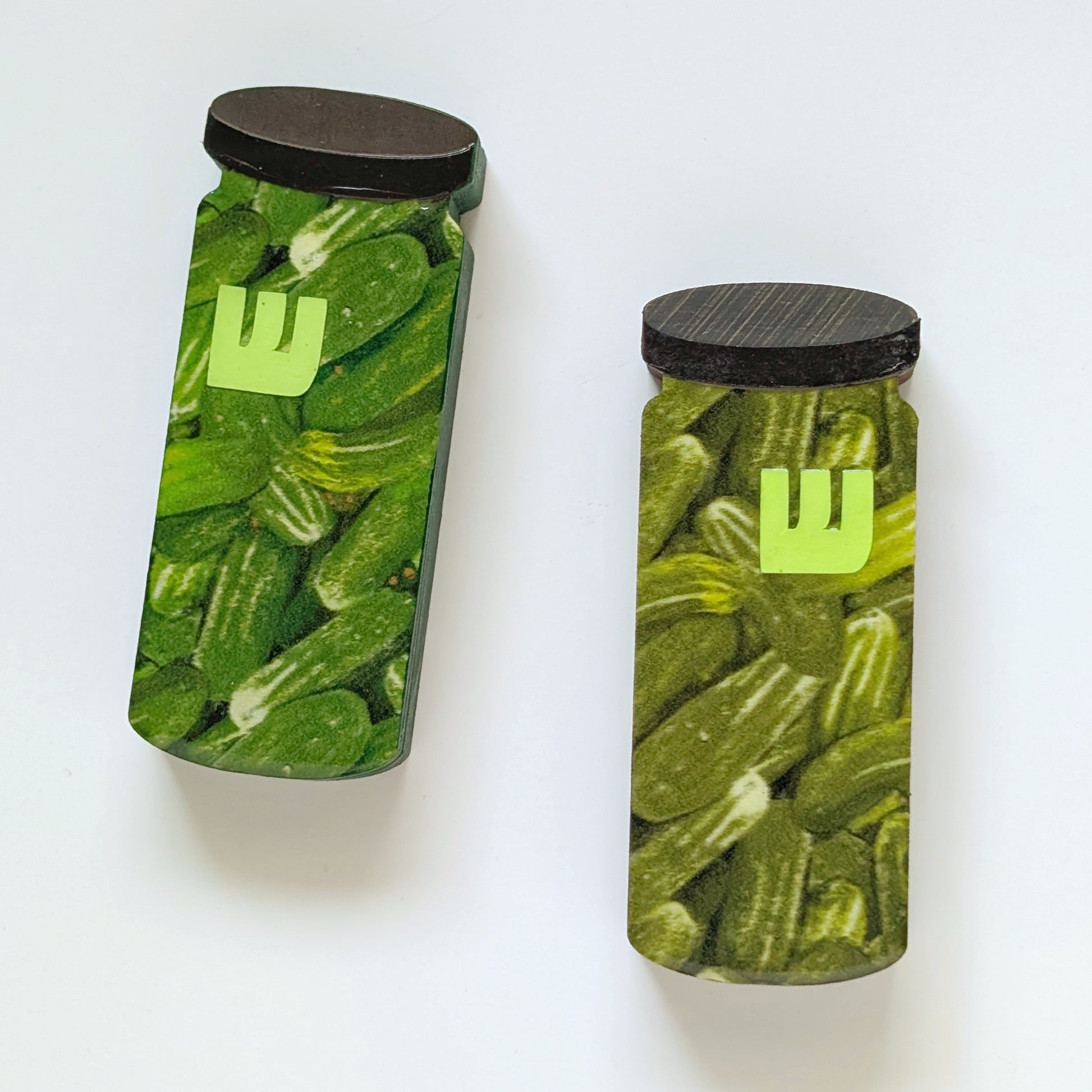 Two mezuzah cases shaped like jars filled with pickles with black lids on a white background