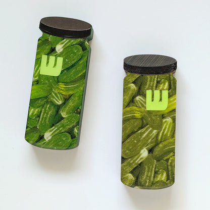 Two mezuzah cases shaped like jars filled with pickles with black lids on a white background