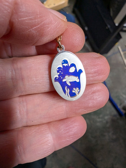 Silver oval pendant with a dark blue hamsa decorated with a turtle and other sea creatures