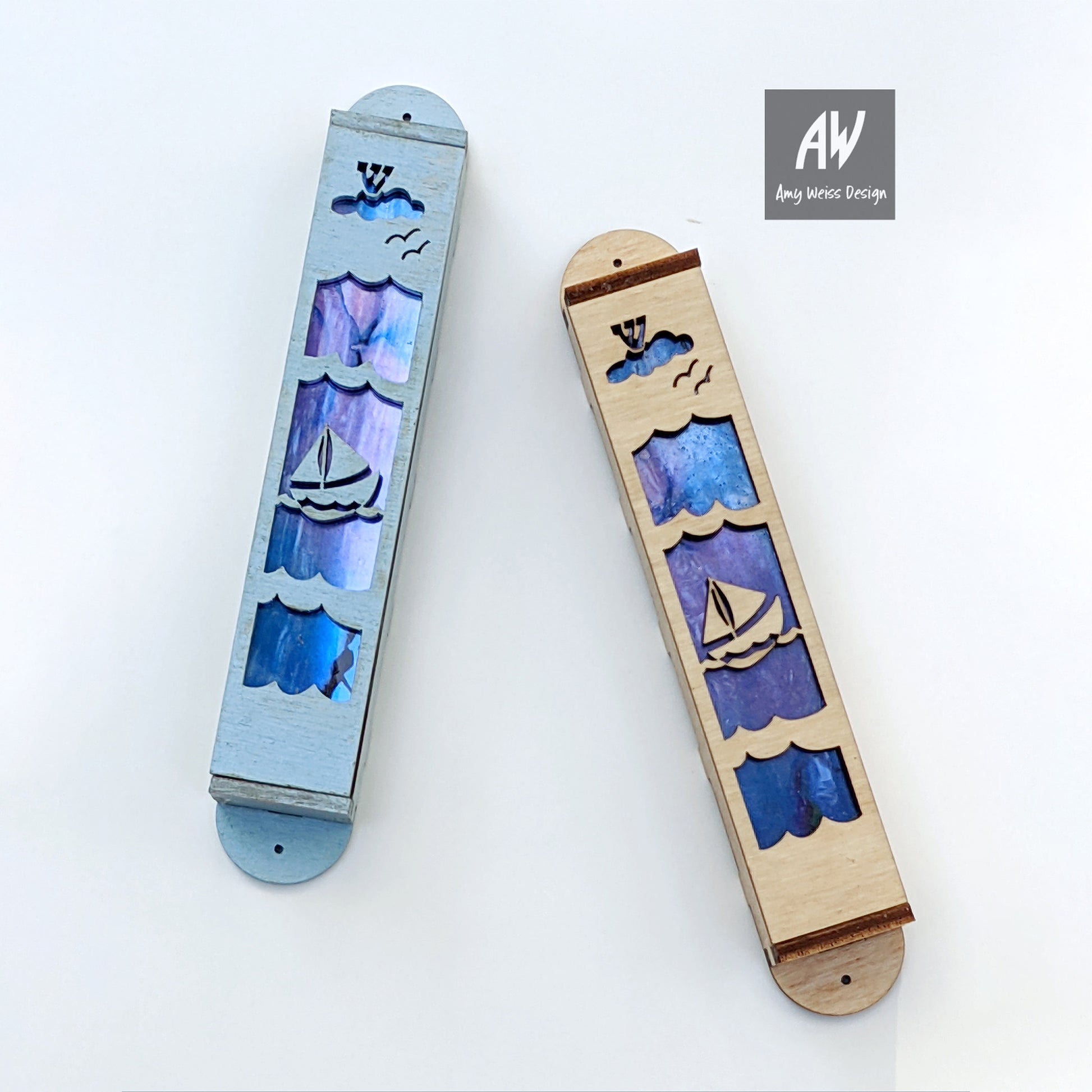 two mezuzahs, one light blur with sailboat, clouds and the letter Shin over blue/purple background and the other mezuzah has the same design with lightly stained wood.